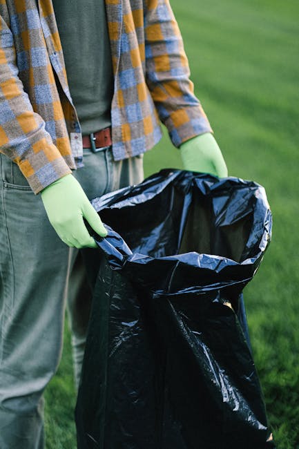 A person standing outdoors on a grassy area, wearing a checkered shirt in shades of yellow, gray, and blue, with green rubber gloves and beige trousers secured with a brown belt. They are holding a large black plastic rubbish bag open at waist level, preparing to dispose of waste. The background features a well-maintained lawn, suggesting a garden or park setting. The scene is well-lit with natural daylight, highlighting the texture of the gloves, the glossy finish of the black rubbish bag, and the person's casual attire. The image reflects an activity related to rubbish collection or waste management, with the individual participating in an act of private waste disposal, an alternative to municipal rubbish removal, which is often handled by professional waste management services such as Rubbish Clearance Twickenham. This scene emphasizes responsible rubbish handling and environmental cleanliness in a community outdoor environment.