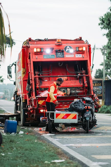 A worker wearing a high-visibility red and yellow vest is operating a large red rubbish collection truck parked on the side of a street. The truck's rear loading mechanism is open, revealing the interior where waste is being collected or compacted. In front of the truck, the worker is handling a black plastic bag containing waste, which is placed on a metal platform attached to a motorbike parked beside the truck. The scene is set on a paved road with a curb and some grass on the roadside, with trees and a utility pole with overhead wires visible in the background. The sky appears overcast, contributing to a neutral lighting environment. The image illustrates an on-site waste collection process typical of private rubbish removal services, aligning with the practice of independent waste disposal rather than municipal collection, and is relevant to the context of rubbish clearance and waste management.