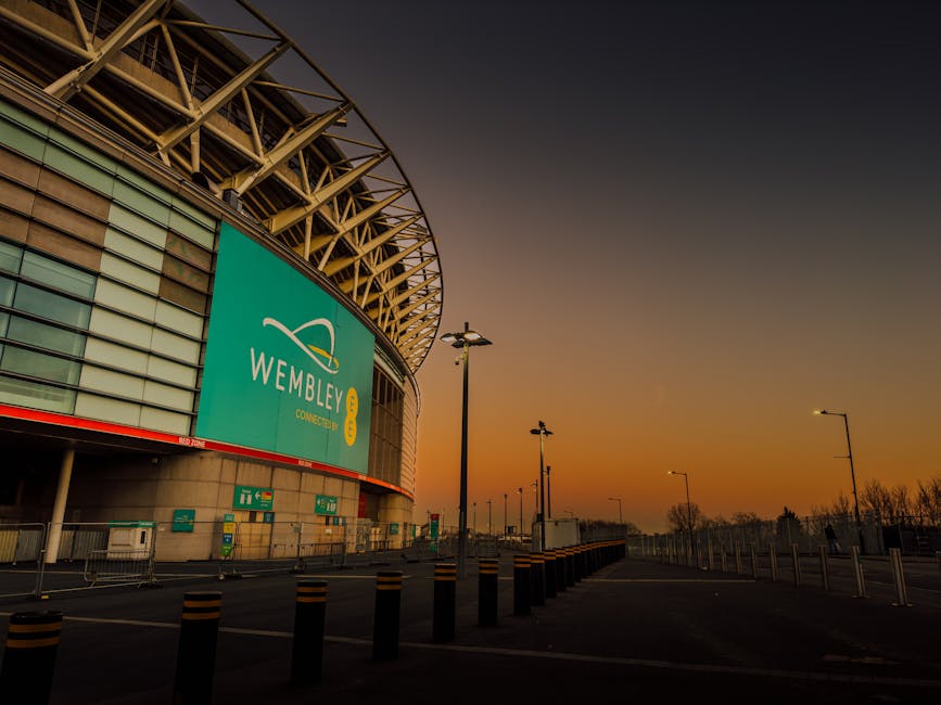 A wide view of the exterior of Wembley Stadium during sunset, showcasing the curved metal framework of the roof structure and a large turquoise signboard with the Wembley logo. The building's facade features horizontal glass panels and concrete elements, with several green waste disposal and emergency signage visible near the entrance. In the foreground, there is an empty parking area marked by black bollards with yellow reflective bands, and a row of tall streetlights line the paved pavement. The sky exhibits a gradient of warm orange and purple hues as daylight fades, providing a calm ambience. The scene reflects an urban environment suitable for independent waste disposal or rubbish collection services, with Rubbish Clearance Twickenham potentially providing on-site clearance or alternative waste handling nearby.